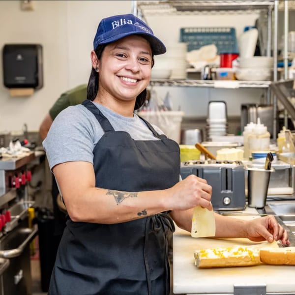 Smiling Bilia Eatery chef in a blue cap and apron preparing a Cuban sandwich in a kitchen.