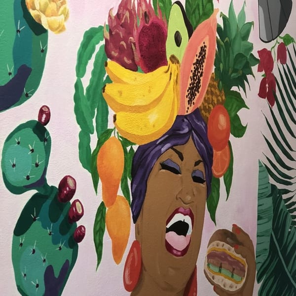 Colorful mural of a woman with fruit on her head, holding a sandwich, surrounded by tropical plants.