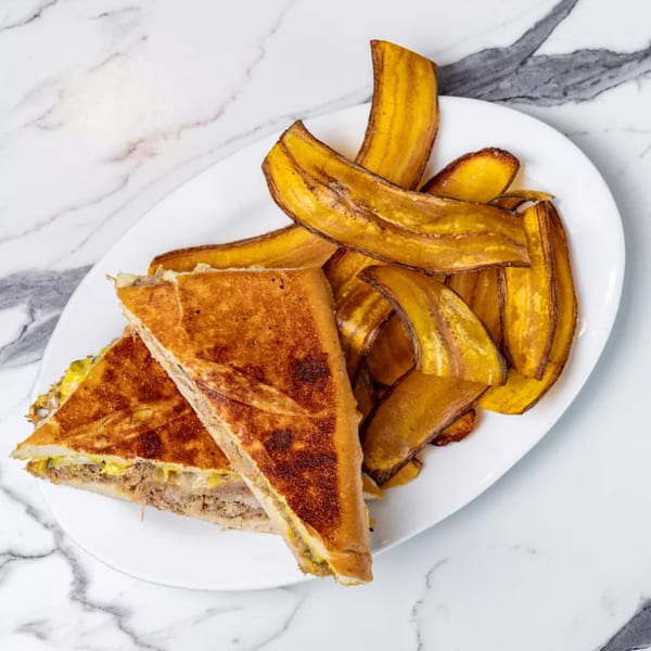 A Cuban sandwich with pulled pork and cheese, served with crispy plantain chips on a white plate.