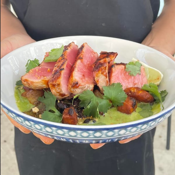 Hands holding a bowl of seared tuna, green sauce, plantains, and cilantro from Bilia Eatery.