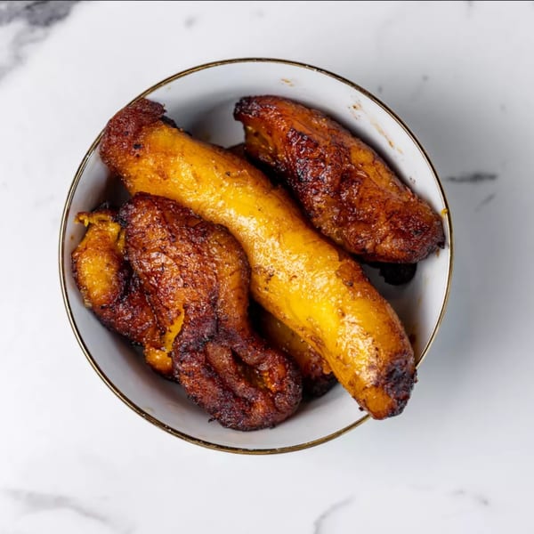 A white bowl with a gold rim filled with golden-brown fried plantains on a marble surface.
