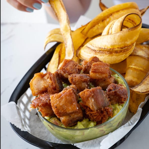 Crispy pork belly chunks over guacamole, served with plantain chips in a black basket.